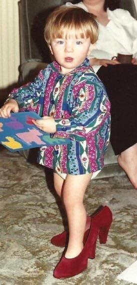 A toddler wearing a woman&#x27;s high-heeled red shoes and a colorful top