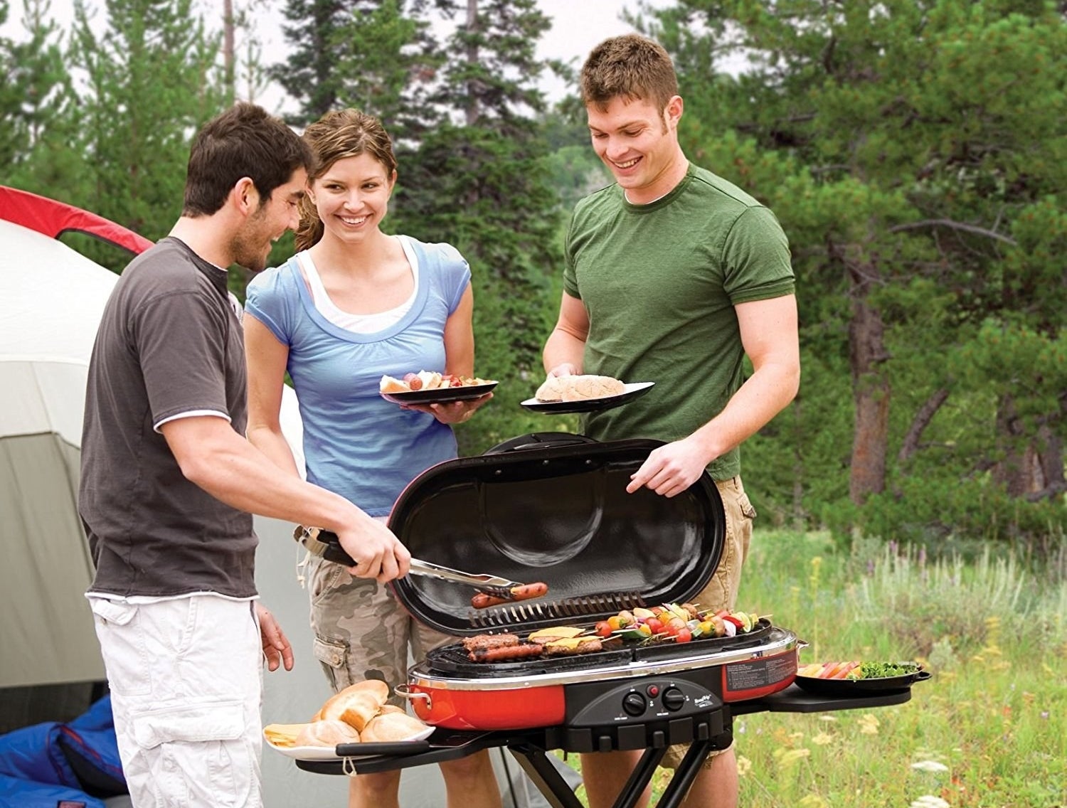 three people using the small portable grill to make kabobs and hot dogs beside of a camping tent