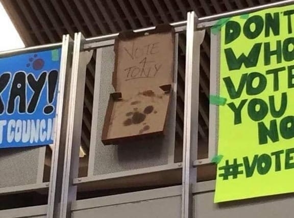 Three posters on a railing: one promotes a student council, a pizza box says &quot;Vote 4 Tony,&quot; and another reads &quot;Don&#x27;t know who to vote for? You now do! #VoteTony.&quot;