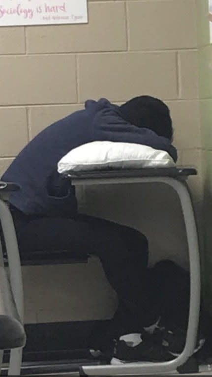 Person in a classroom resting their head on a pillow placed on a desk. Poster on the wall reads, &quot;Sociology is hard.&quot; No text on the person&#x27;s clothing is visible