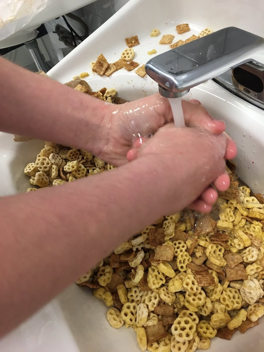 Hands are being washed under a running faucet with various dry cereals scattered in the sink