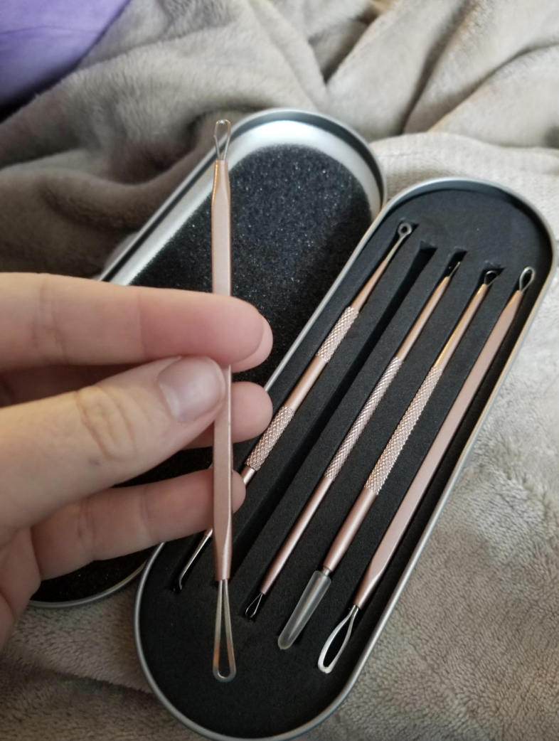 A silver tin (shaped like an eyeglass case) is black on the inside and has five skinny slots where the blackhead remover tools are sitting. A hand is holding one of the tools which has a looped end.