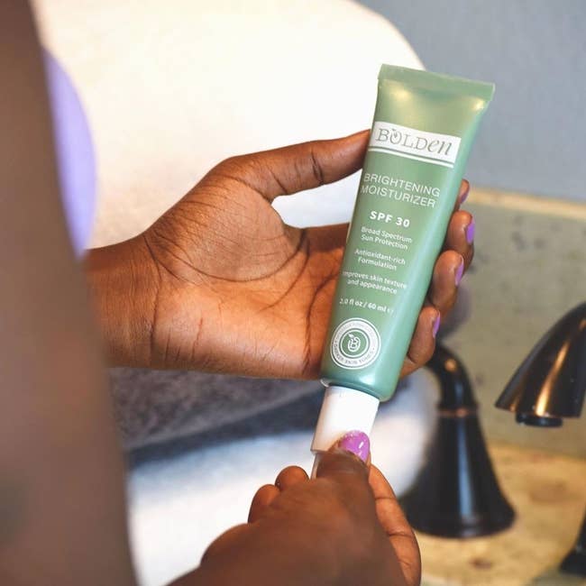 Model opens green and white tube of Bolden's SPF 30 Brightening Moisturizer