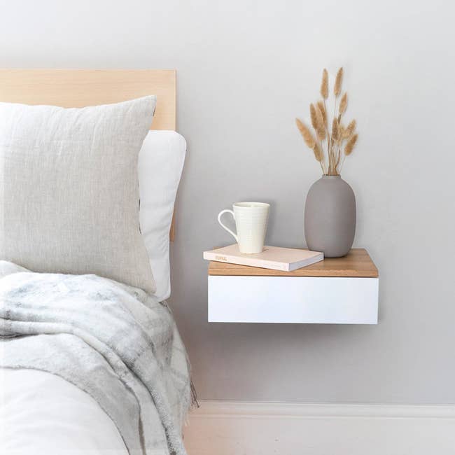 Rectangle white wood drawer with a light brown wood top attached to the wall next to the bed with a book, coffee cup, and vase on it