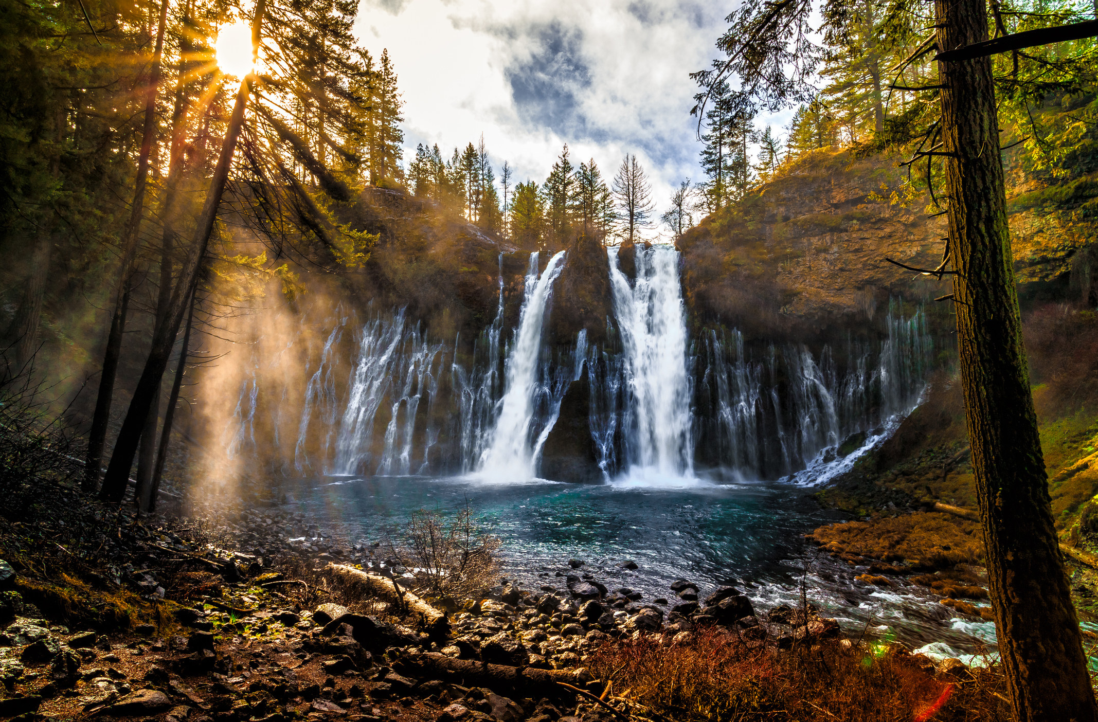 A cascading waterfall with the sun and trees nearby.