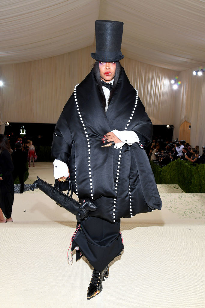 Erykah Badu wears a long dark oversized cape-like coat and a matching top hat