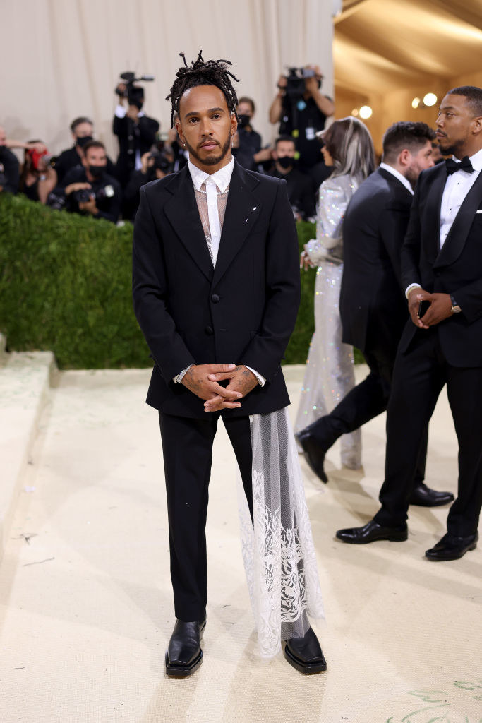 Lewis Hamilton wears a light colored button up shirt that has a long lace train on the hem under a dark suit jacket with matching slacks