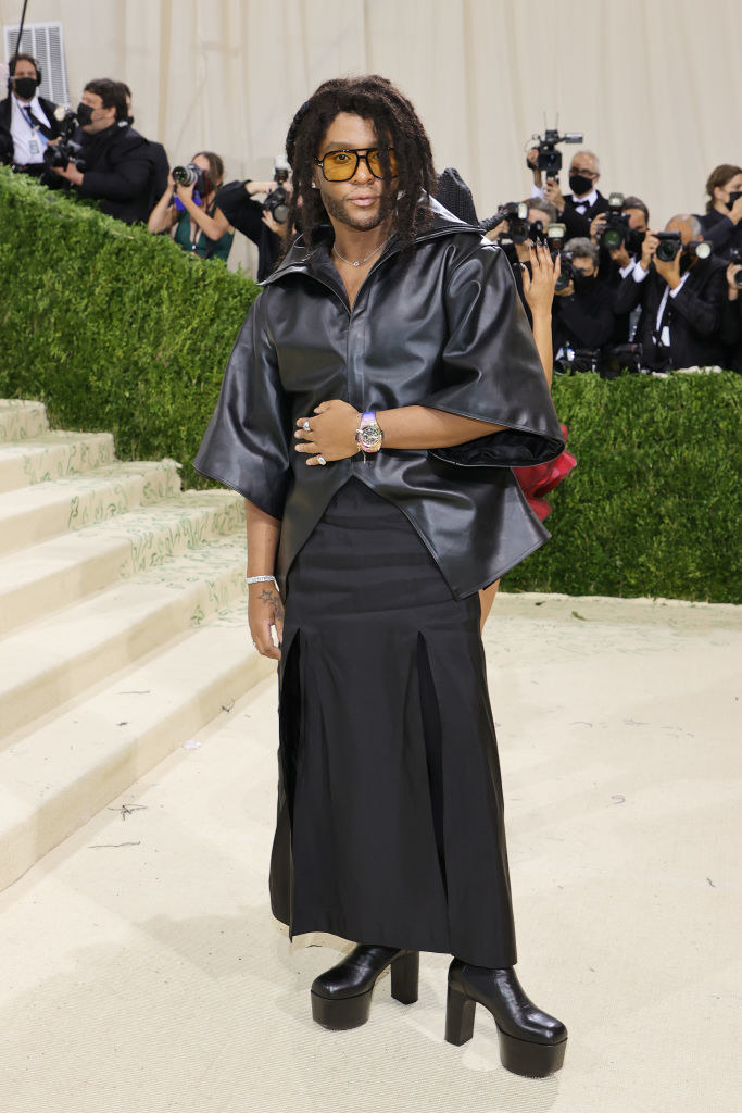 Law Roach wears a long dark skirt, matching colored platform boots, and a short sleeve leather blouse