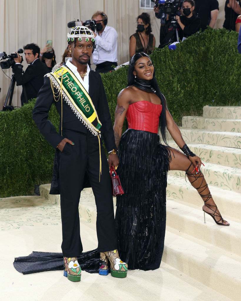 Edvin Thompson wears a dark suit with a bright sparkly slash around his torso and a matching beaded head piece
