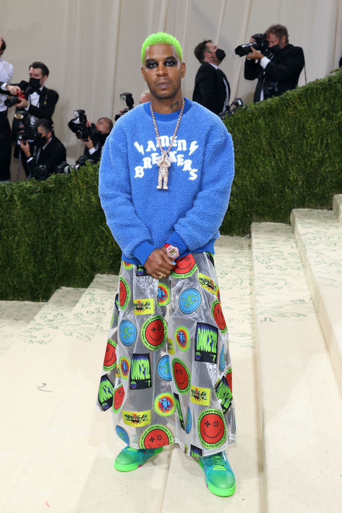 Kid Cudi wears a fuzzy brightly colored sweatshirt over a long skirt with neon colored patches on it