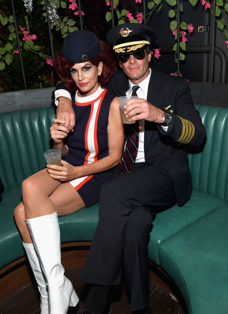 Cindy Crawford wears a short sleeveless dark dress with bright knee length boots and Rande Gerber wears a dark suit and matching sunglasses