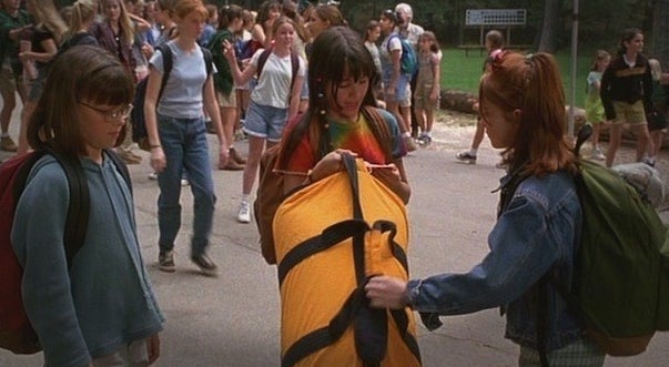 A girl in a tie-dye shirt holds a yellow duffle. A red haired girl reaches for it