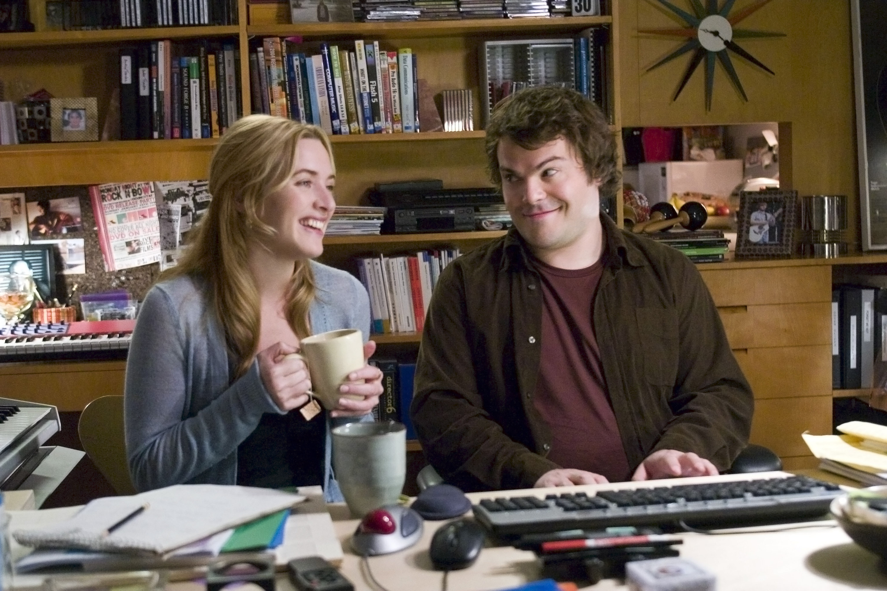 A man and woman sit in an office as he plays a keyboard piano