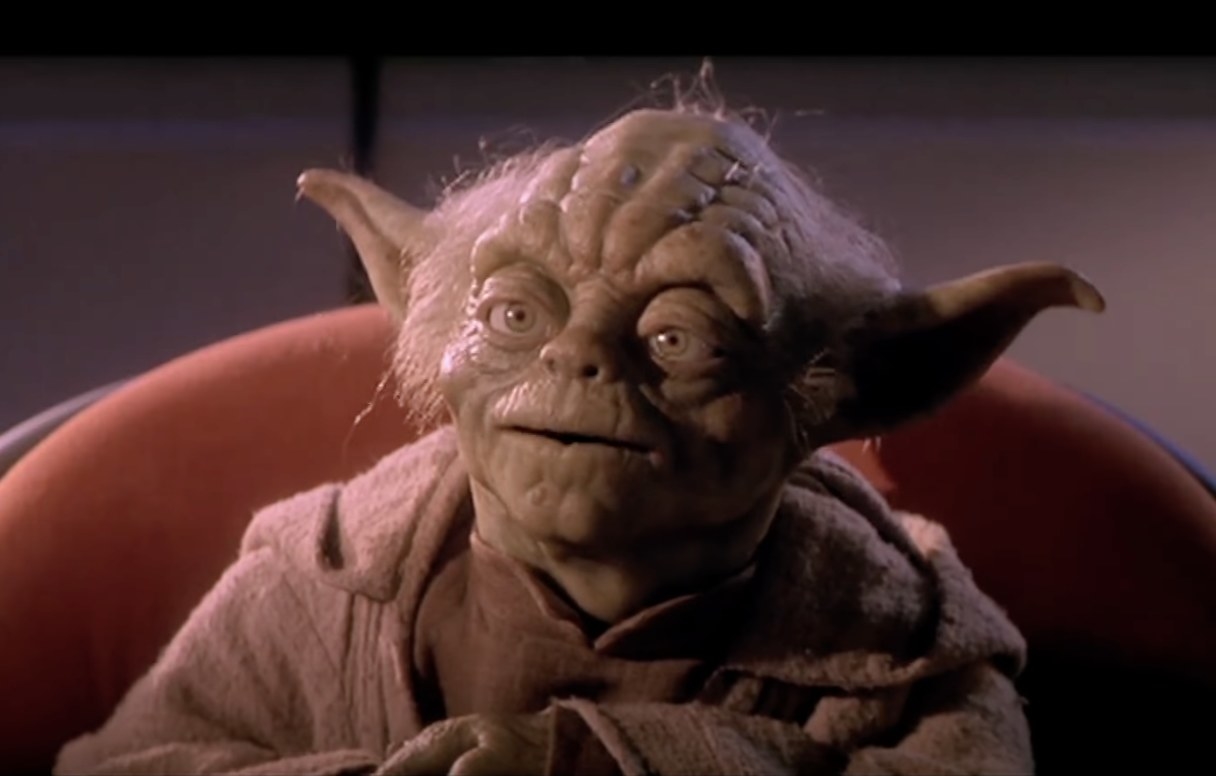 Yoda, a character from Star Wars, sits on a chair wearing a brown robe, with a wise and calm expression