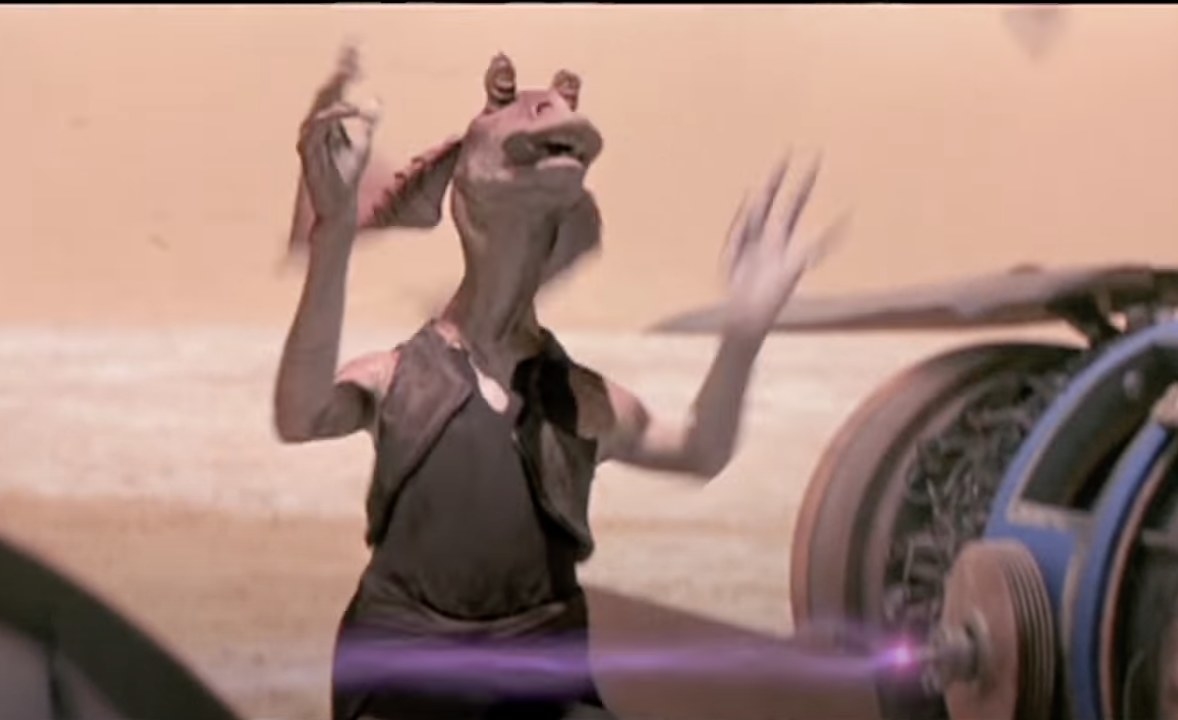 Jar Jar Binks stands with raised arms near a pod racer in a desert setting from a Star Wars film