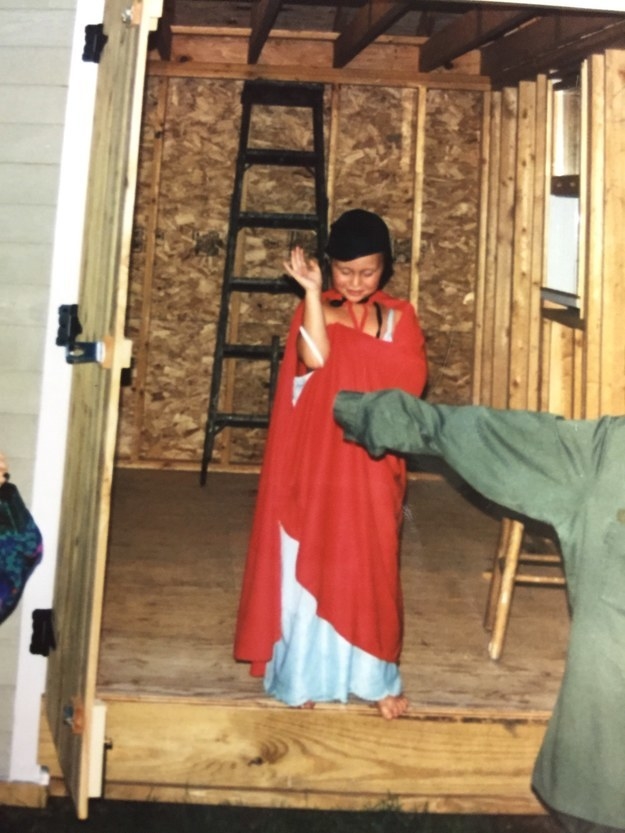 A child with their eyes closed and waving wears a long cape and gown and a helmet hat