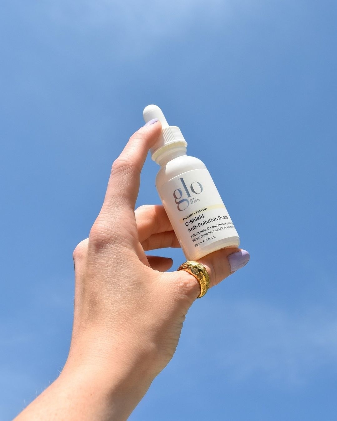 A hand with gold ring on its thumb holds a small white bottle with dropper to a blue sky.