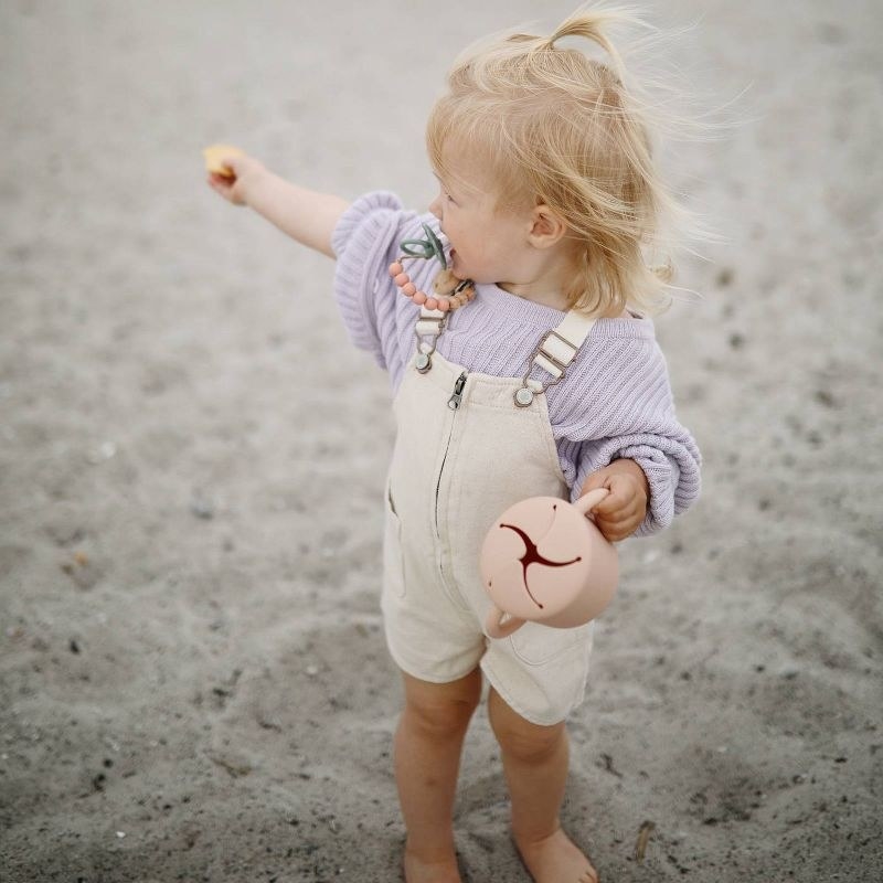 Young child on a sandy beach wearing a knit sweater and overalls, holding a snack and a sippy cup