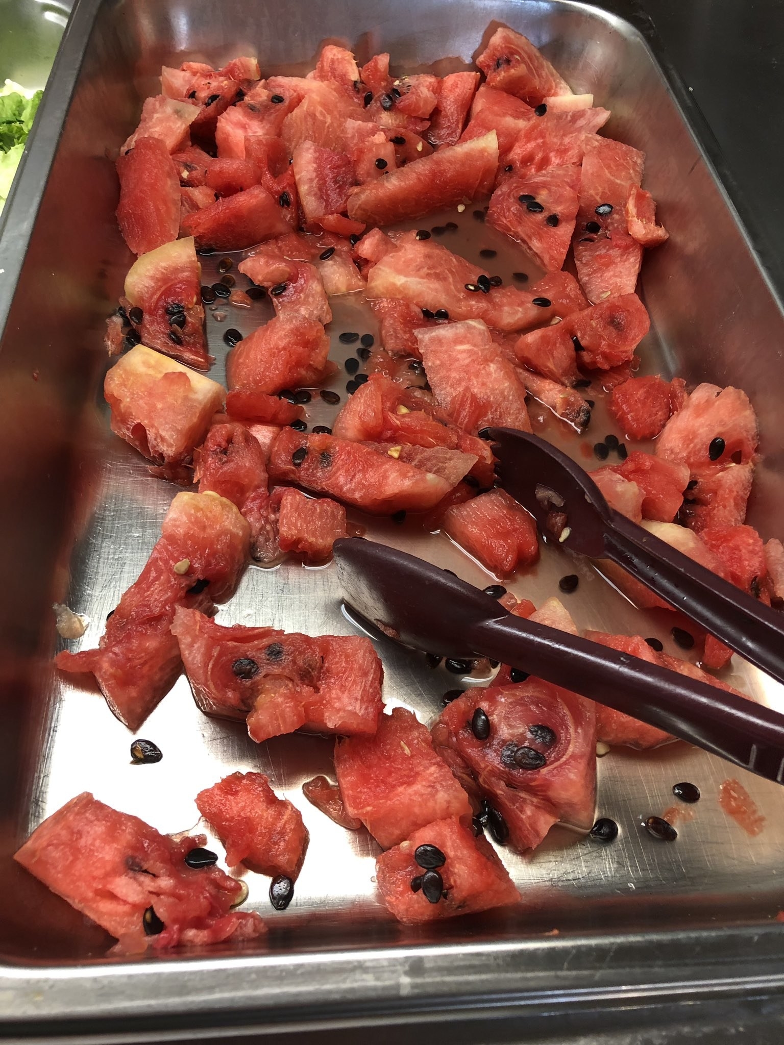 A stainless steel tray filled with chunks of watermelon and scattered black seeds. Red tongs are placed within the tray for serving