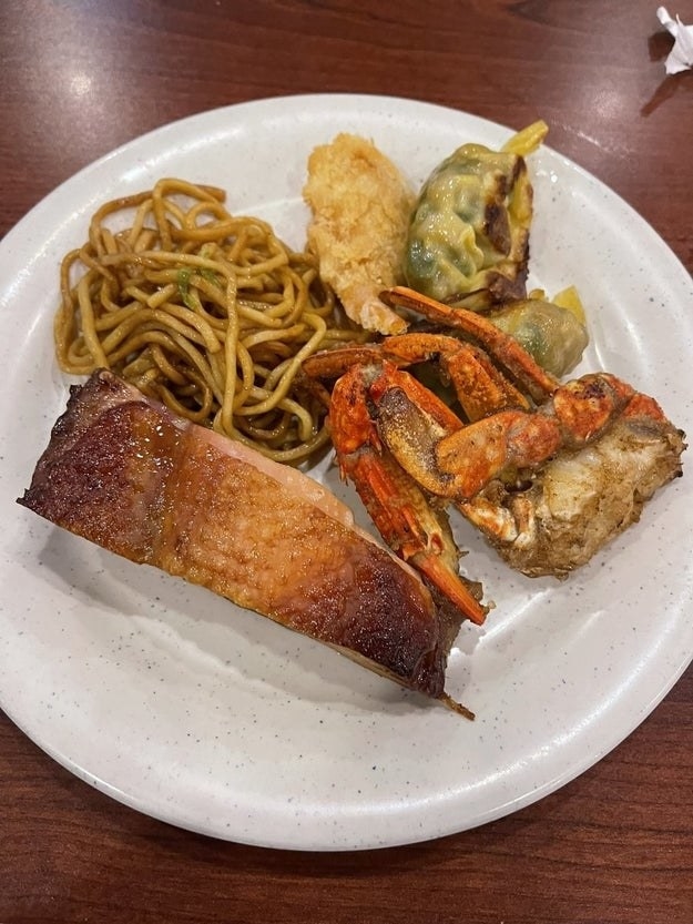 A plate with assorted food items including noodles, a piece of meat, breaded shrimp, crab legs, and a stuffed green pepper