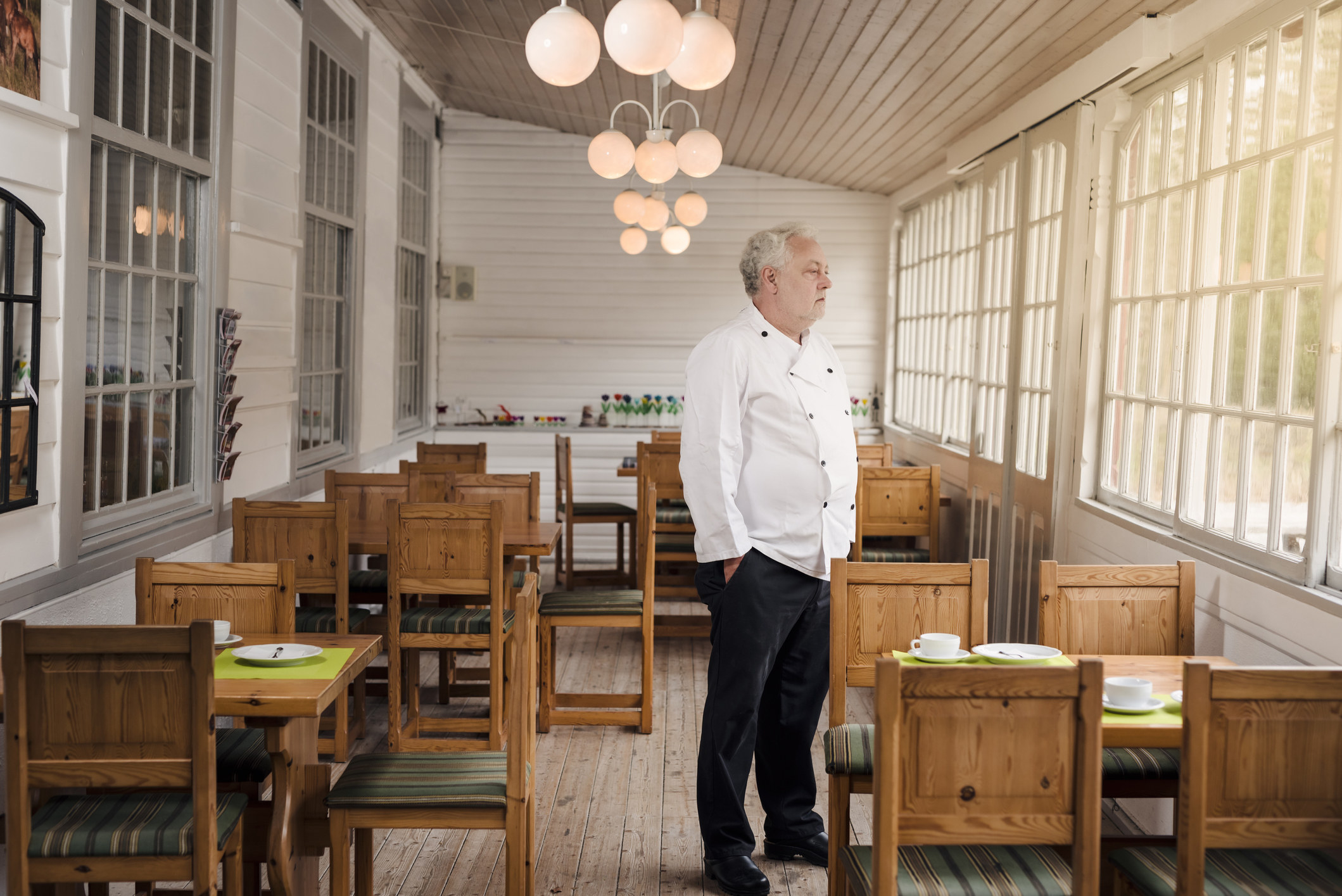 A man in a chef&#x27;s uniform stands in an empty, rustic dining area with wooden chairs and tables, looking out the window