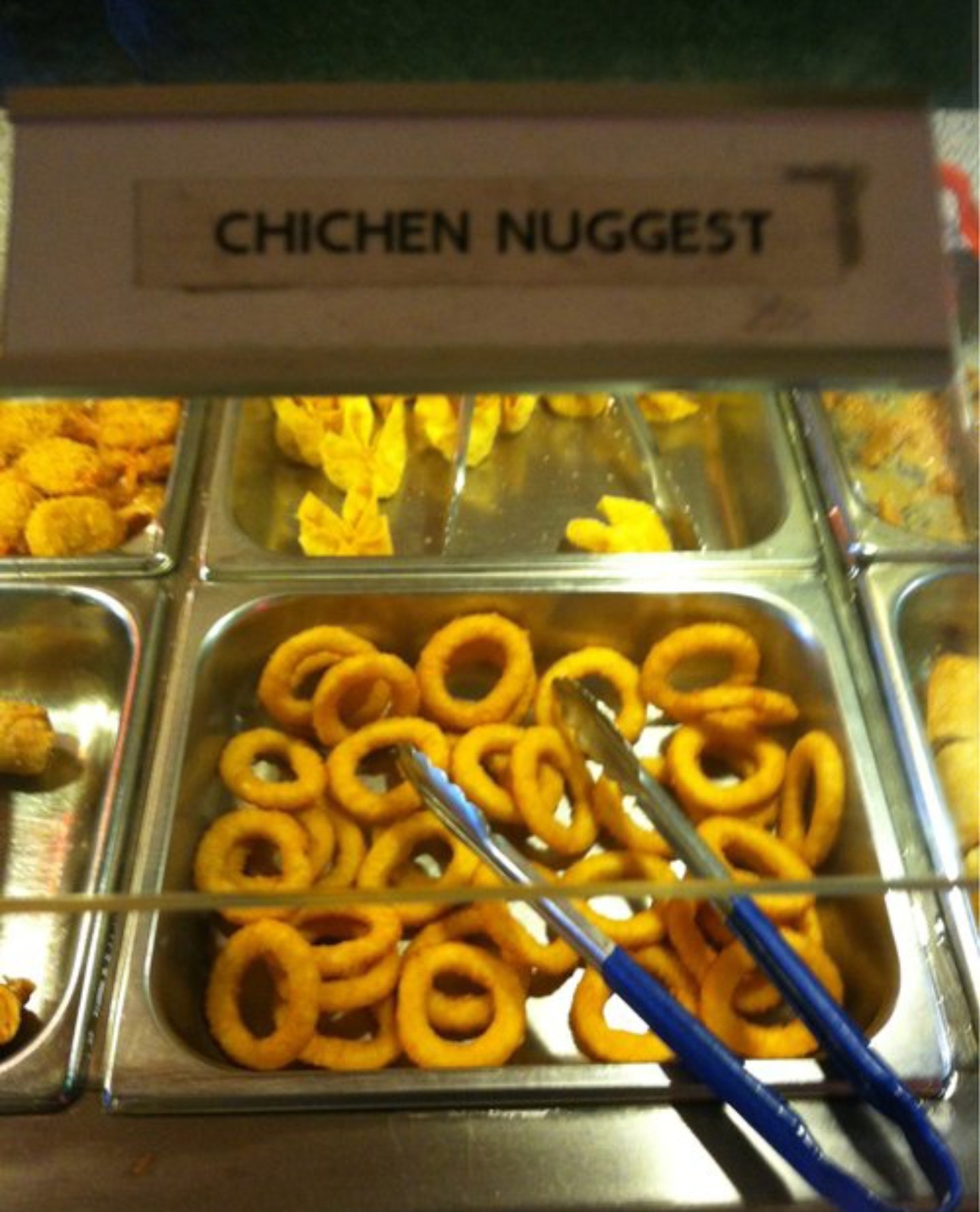 A buffet tray filled with onion rings under a sign that reads &quot;CHICHEN NUGGEST.&quot;