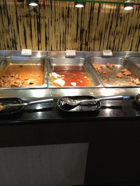 Partially empty buffet trays with various food remnants under a glass shield. Labels on the trays are not readable from this angle