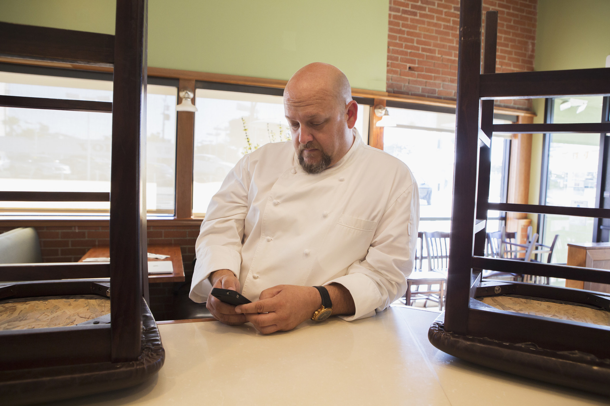 A chef stands in a restaurant, looking at his phone. Chairs are flipped on the tables around him. His expression and posture suggest he is reading something important