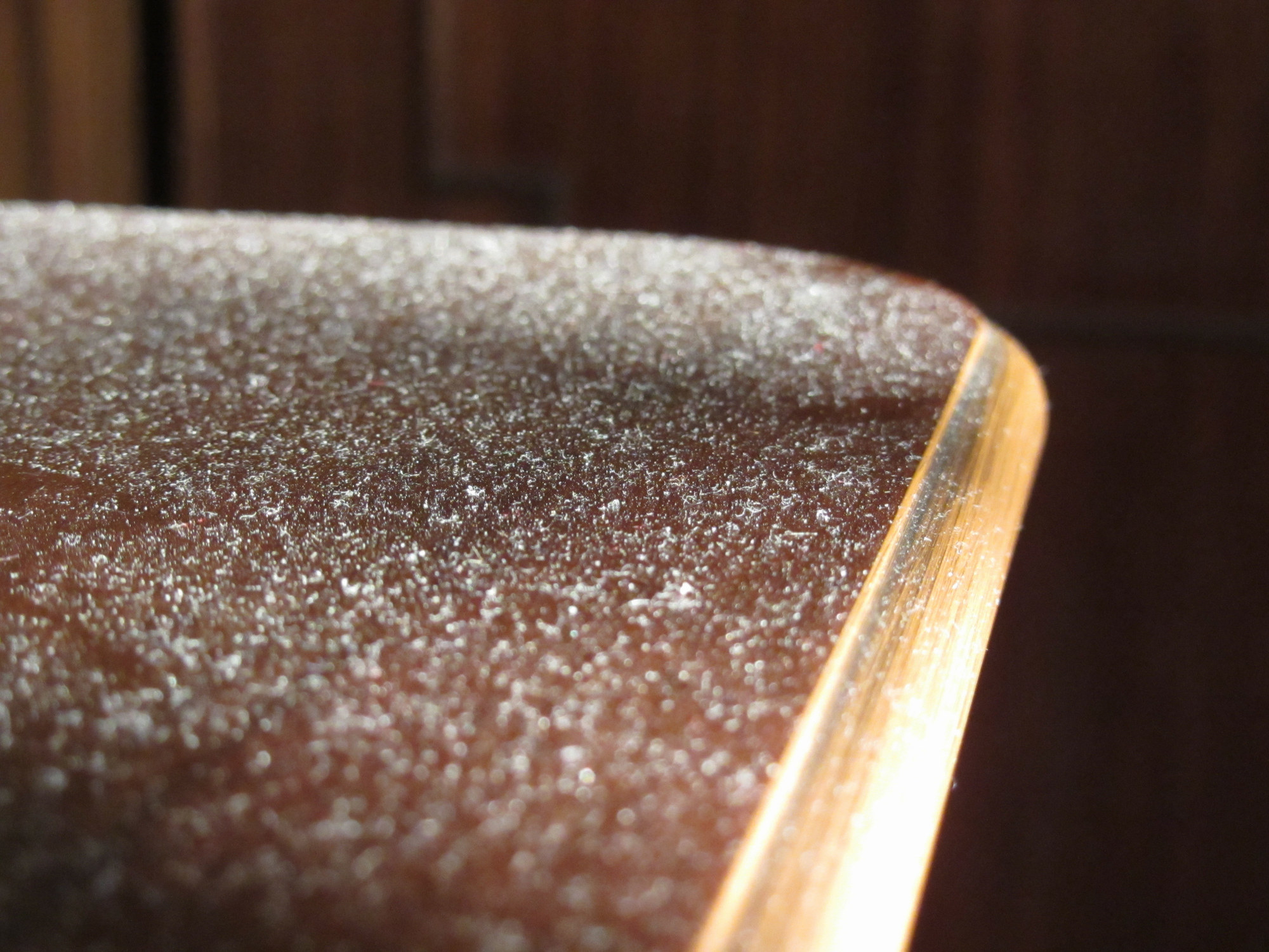 A close-up of a dusty wooden surface corner, highlighting the accumulation of dust particles. No people or significant color details visible