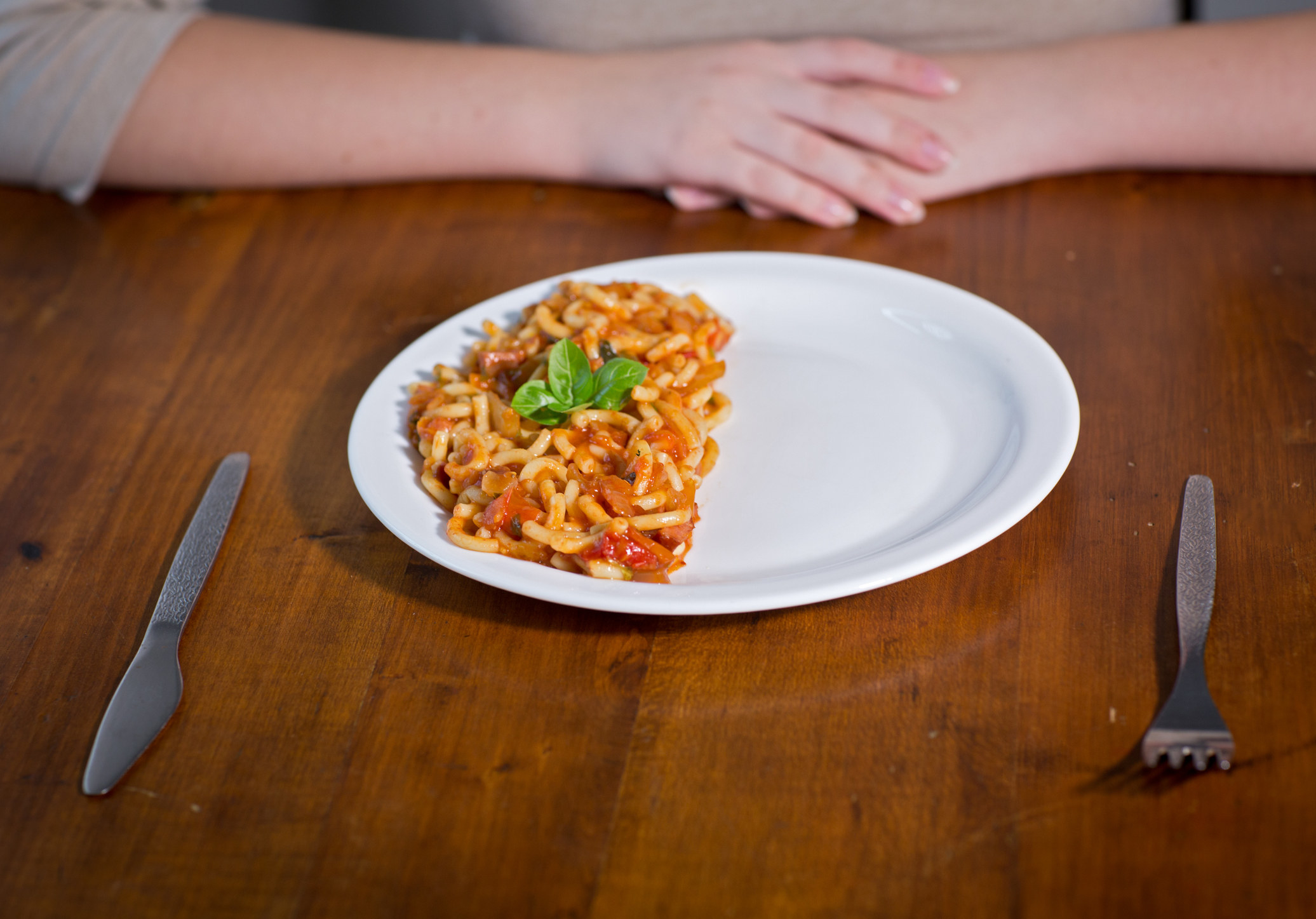 Person seated with hands folded, with a half empty plate of pasta in front of them, knife and fork on either side of the plate