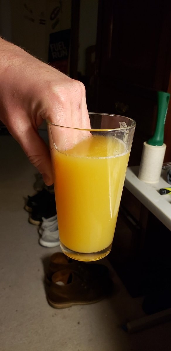 A hand holding a glass of orange juice, with fingers dipped into the drink. The background includes household items and a pair of shoes on the floor