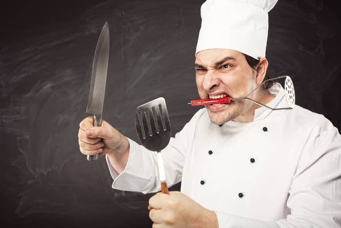 A chef in a traditional white uniform and hat energetically holds a knife, a spatula, and a masher, with a spatula handle in his mouth