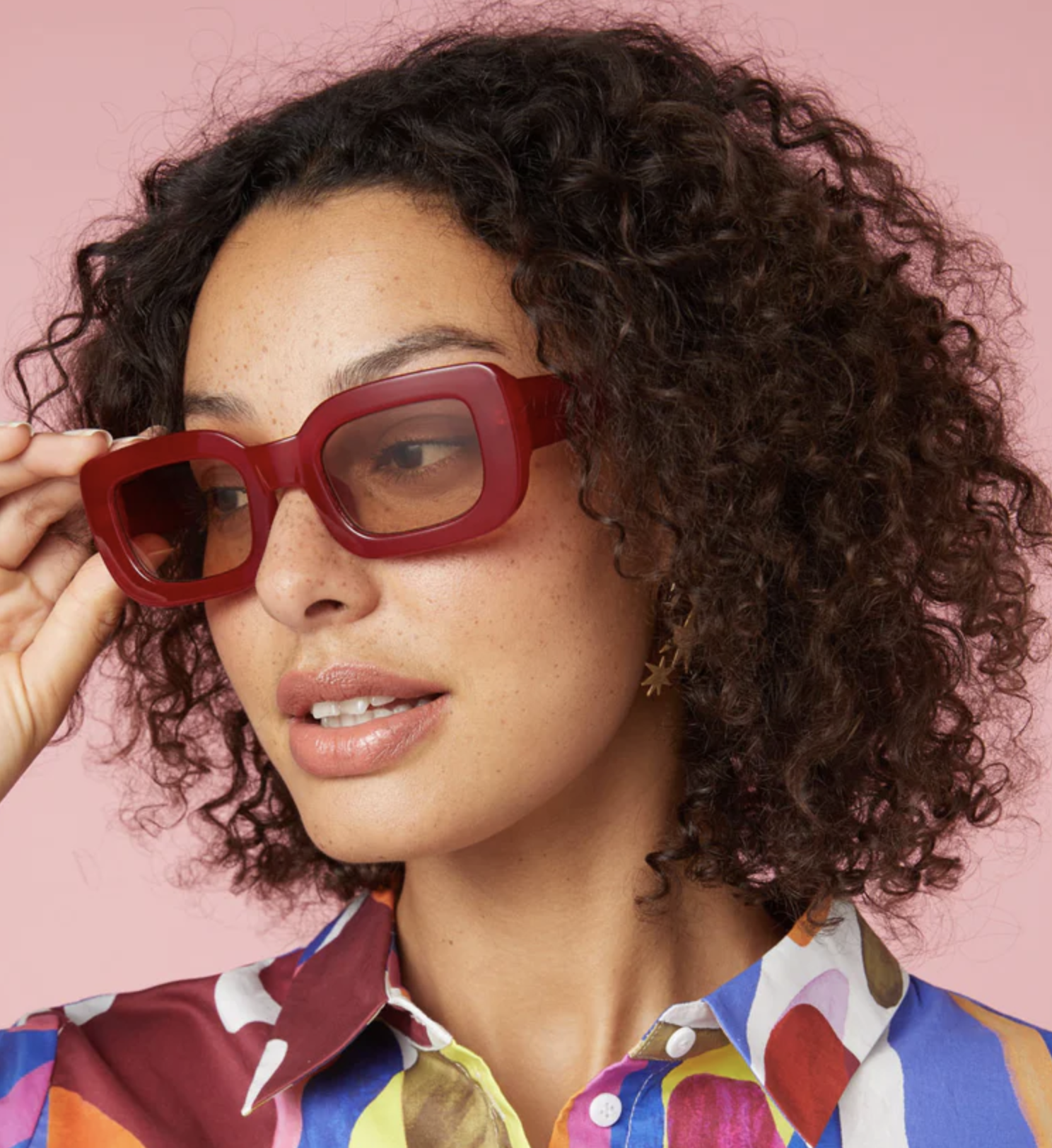 Woman with curly hair, adjusting oversized glasses, wearing a printed shirt