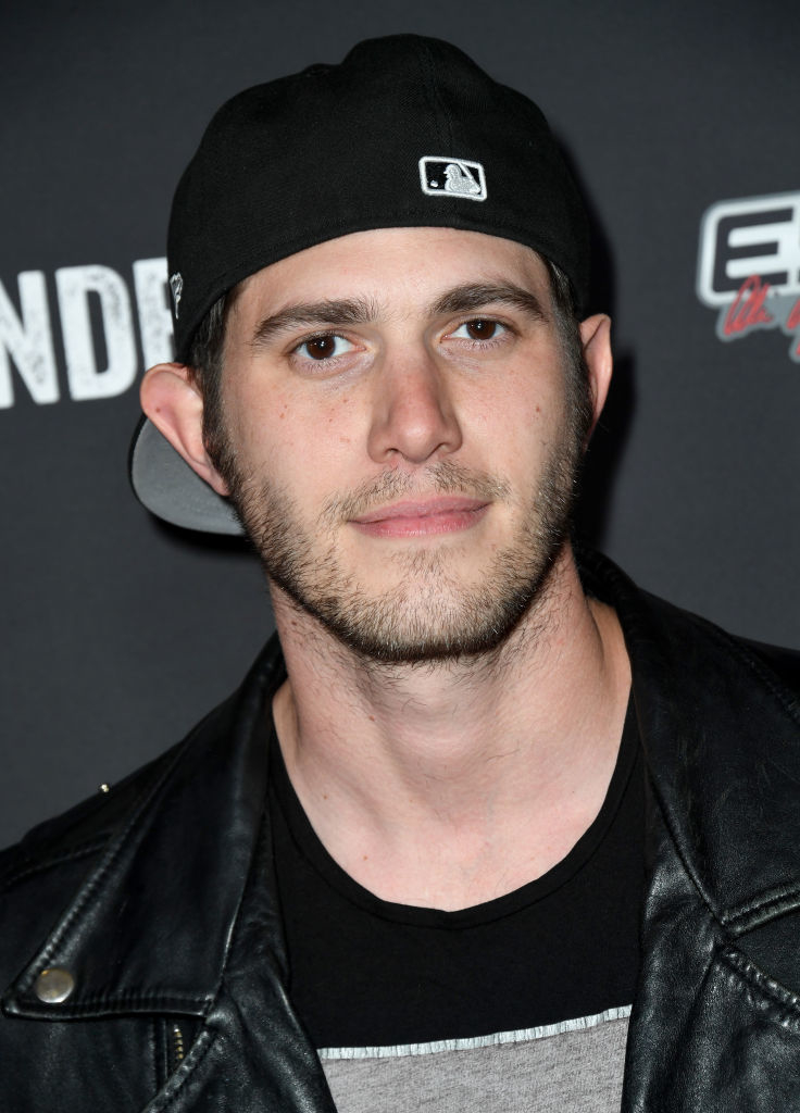 A man in a black baseball cap and leather jacket at an event