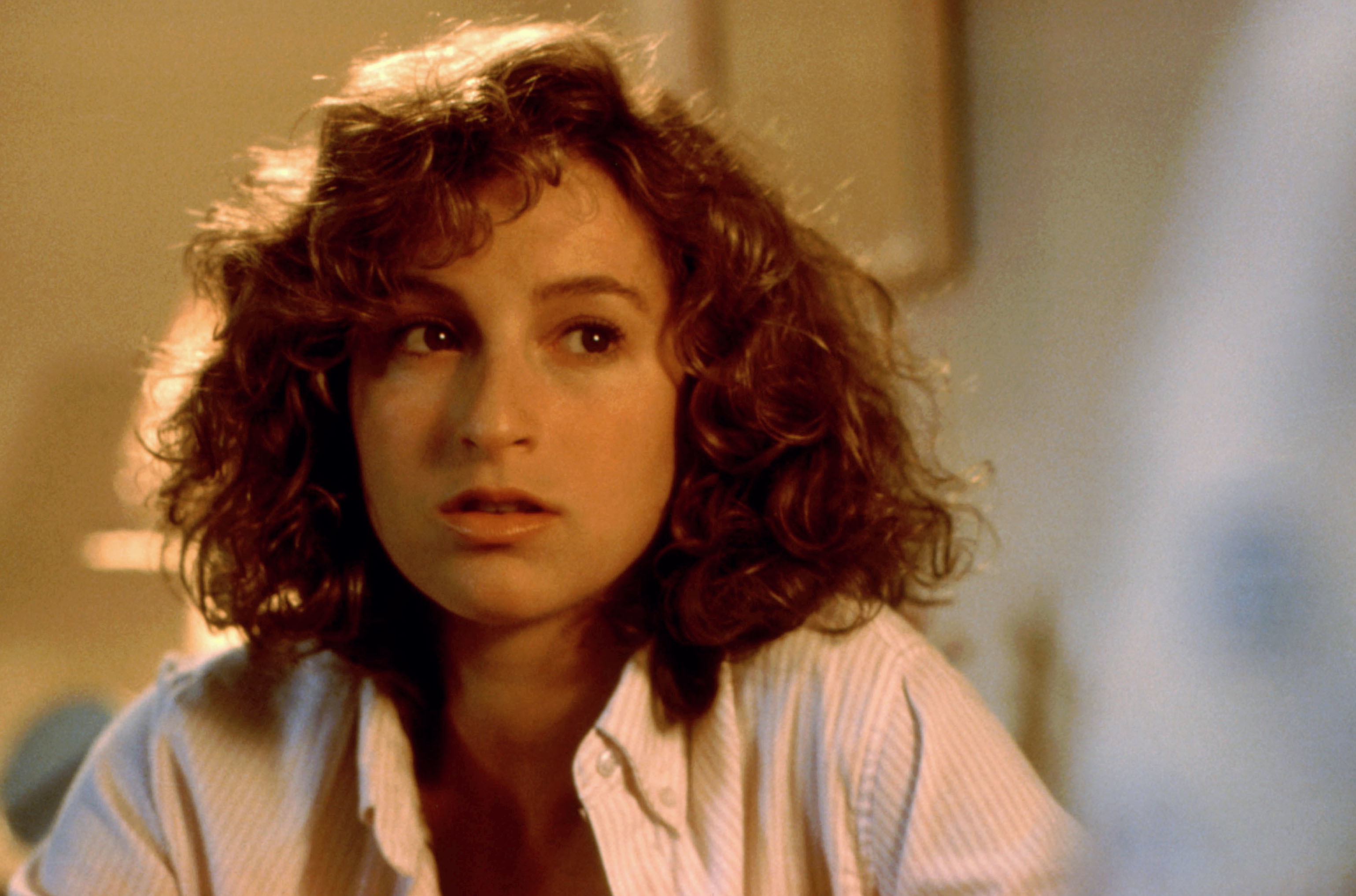 A young woman with curly hair looks to the side, appearing thoughtful. She wears a casual, partially-buttoned shirt