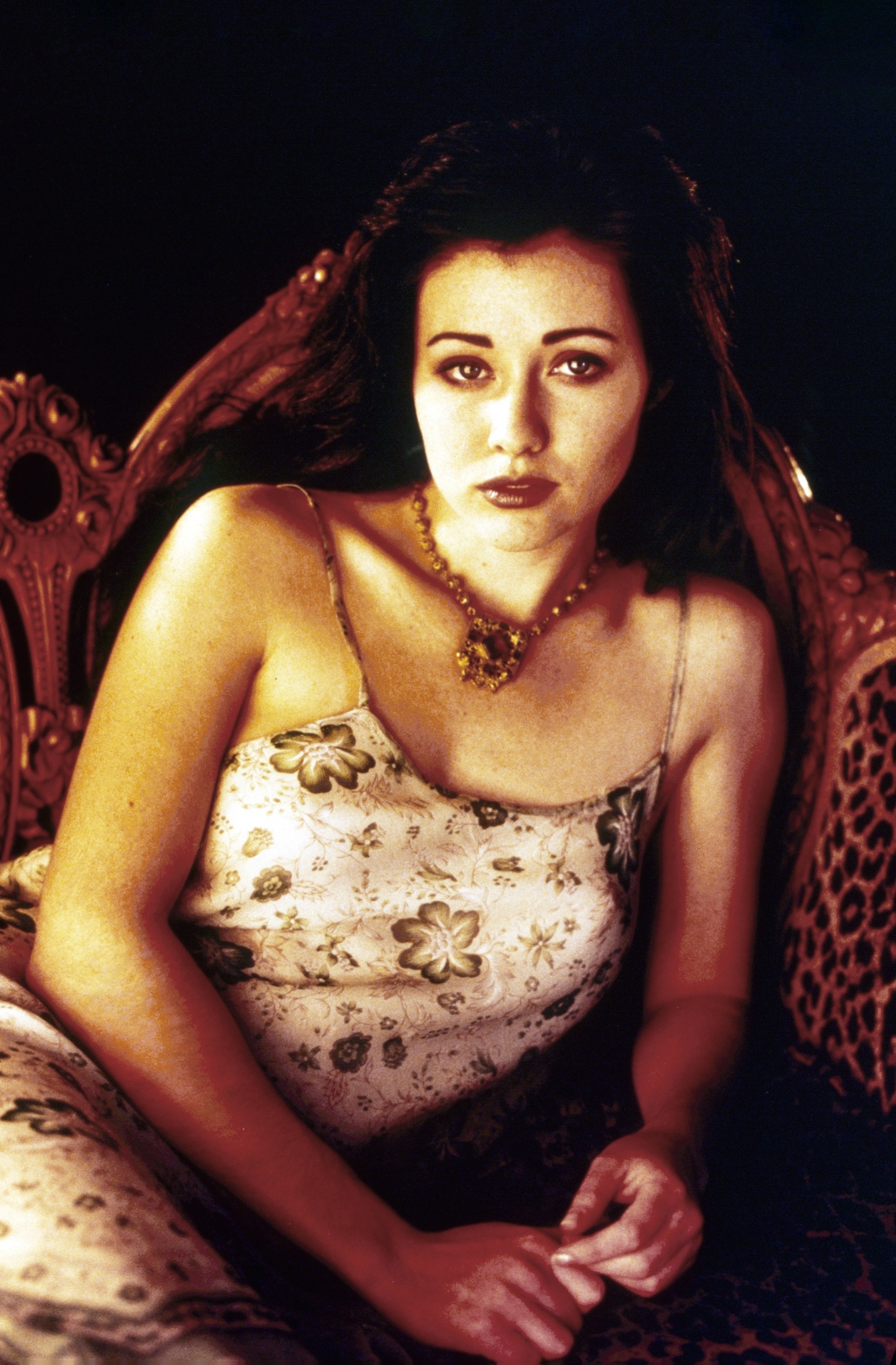 Shannen Doherty poses on a vintage couch wearing a floral spaghetti-strap dress and a necklace
