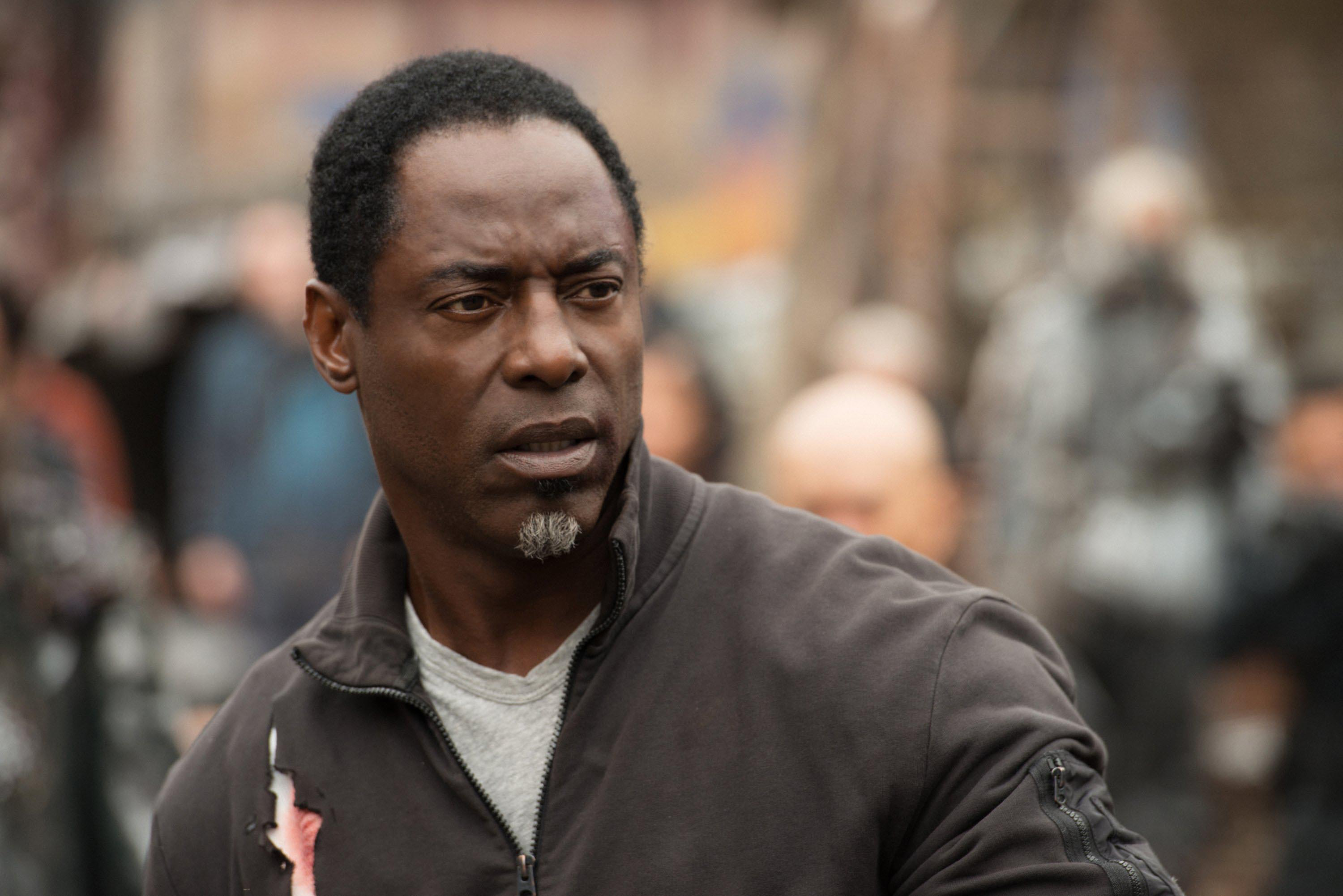 Isaiah Washington wearing a casual jacket, looking concerned in an outdoor setting. Background shows figures and a blurred environment