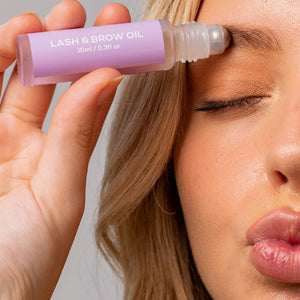 Person applying lash and brow oil from a labeled bottle close to their eyes, demonstrating product use