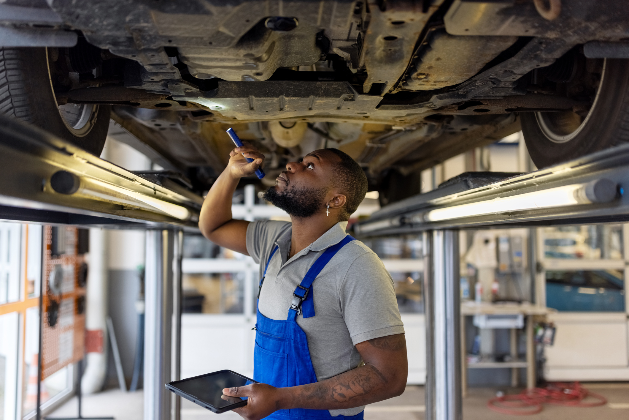 Mechanic in overalls using flashlight to inspect undercarriage of vehicle, holding tablet