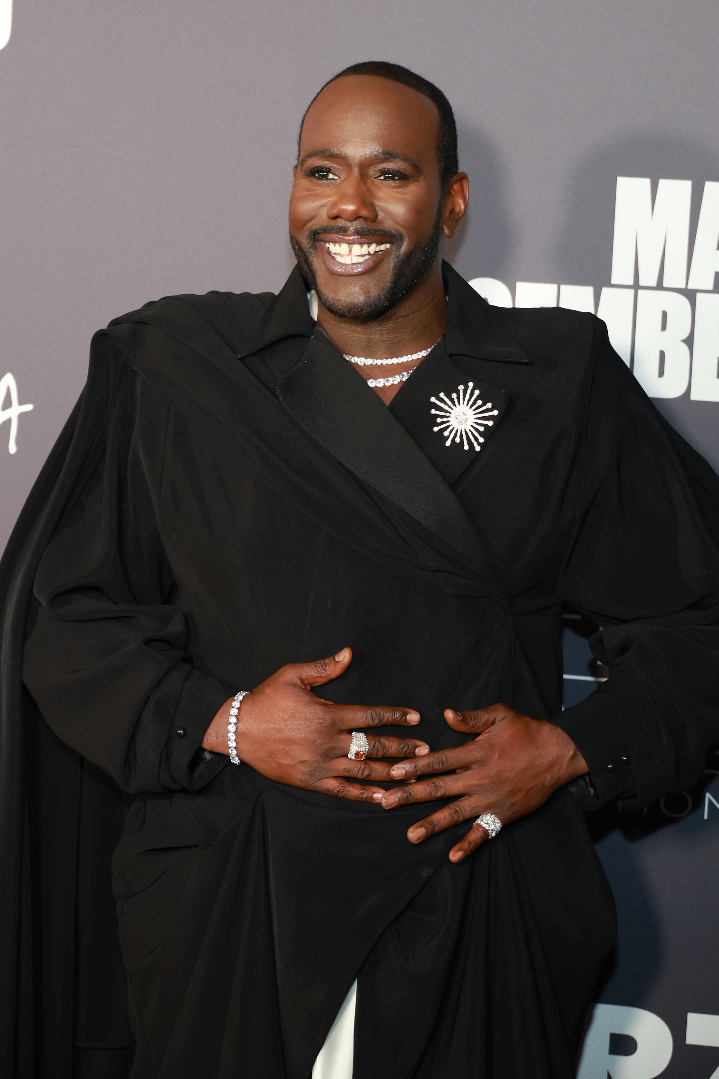 A closeup of Nicco wearing a large, distinctive brooch on the lapel of a wrapped suit smiling at an event