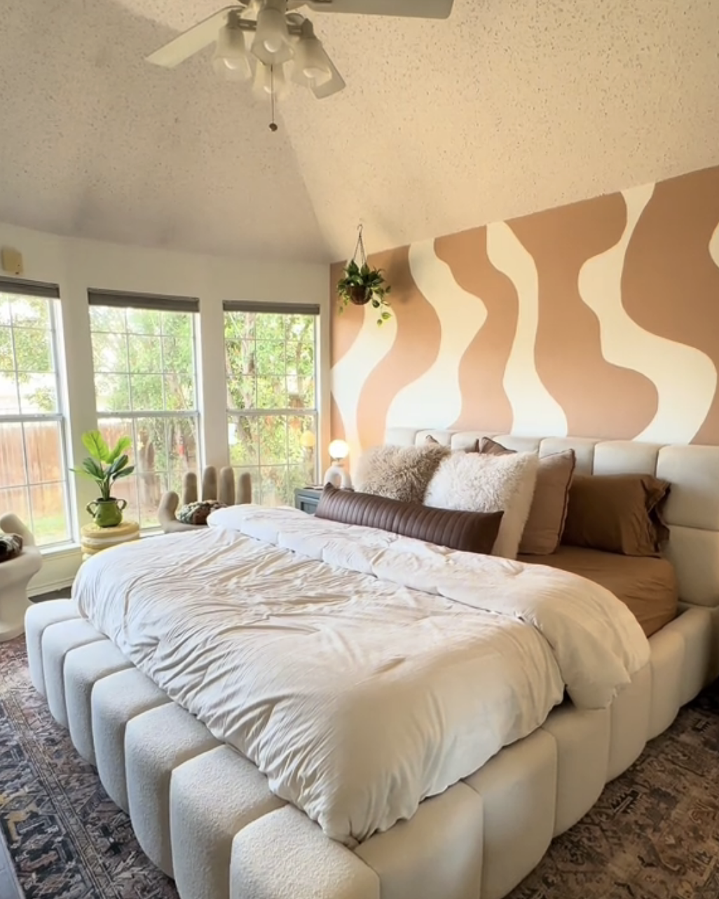 A bedroom with a large bed with pillows, patterned wall, ceiling fan, and windows offering natural light