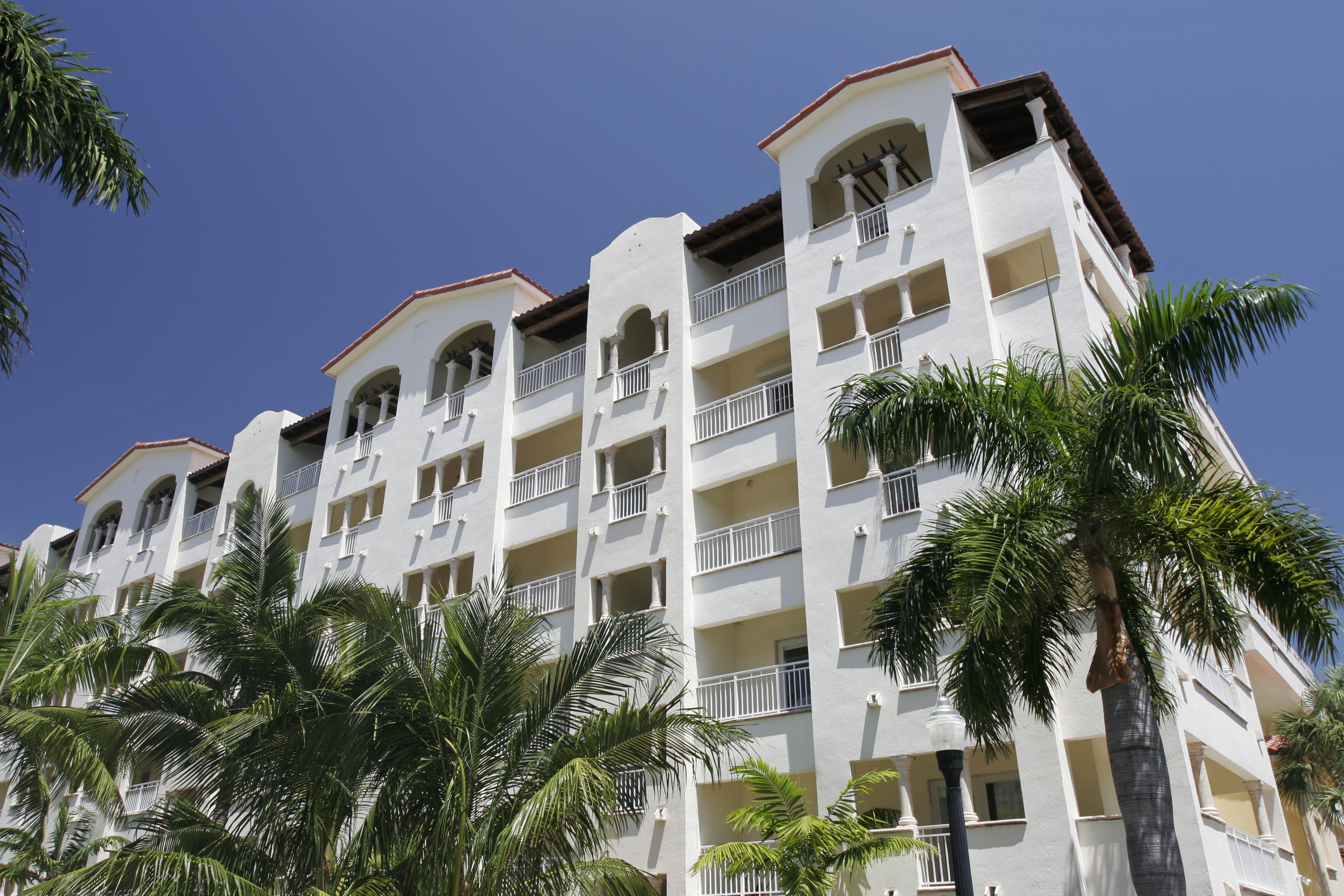 A sunny view of a multi-story residential building with a white facade and red trim, surrounded by palm trees