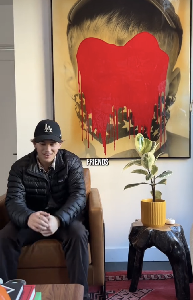 Deacon in cap and jacket seated by artwork with dripping heart design