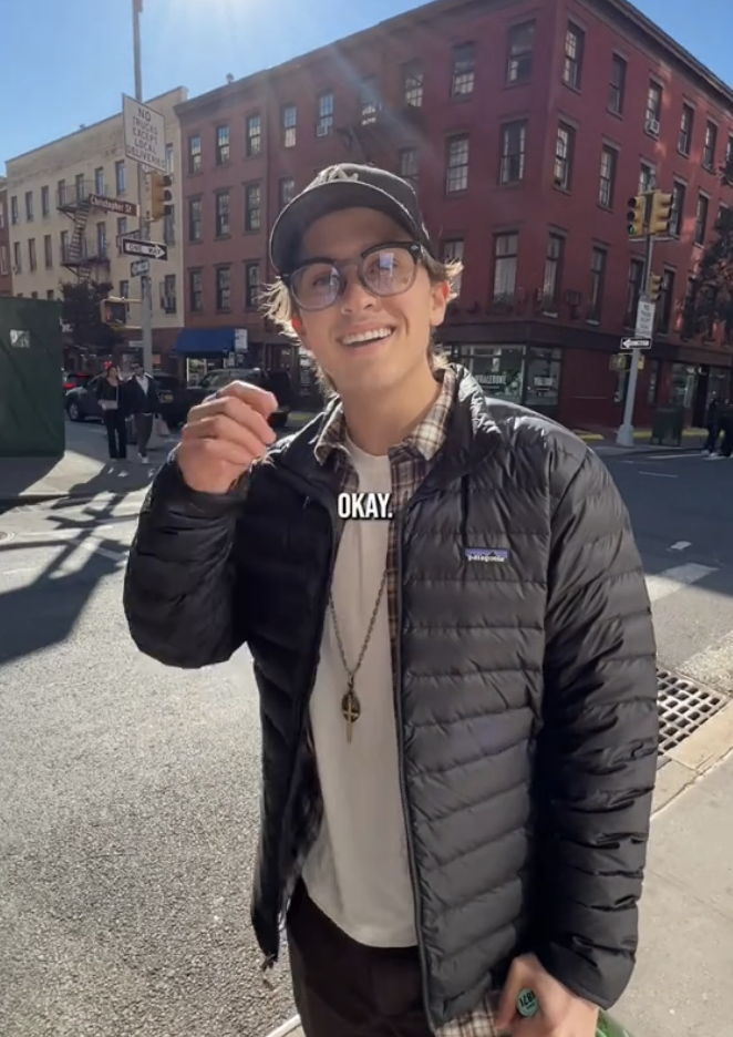 Deacon in a jacket and cap smiling on a city street, gesturing with his hand