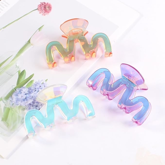 Three iridescent claw-style hair clips on a white surface, next to flowers