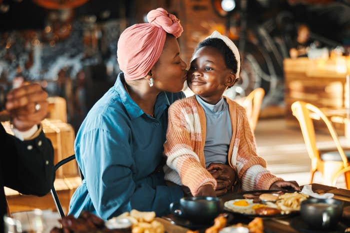 Woman kissing child on the cheek at a table with food, sharing a tender moment