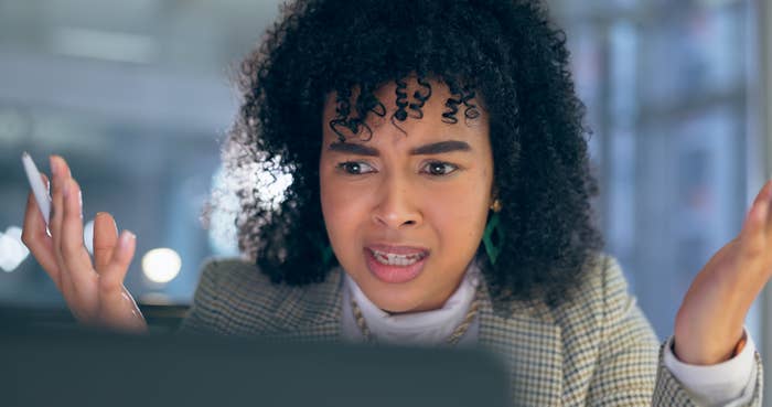 Woman appears frustrated or confused looking at a computer screen, hands raised slightly. She&#x27;s wearing a blazer