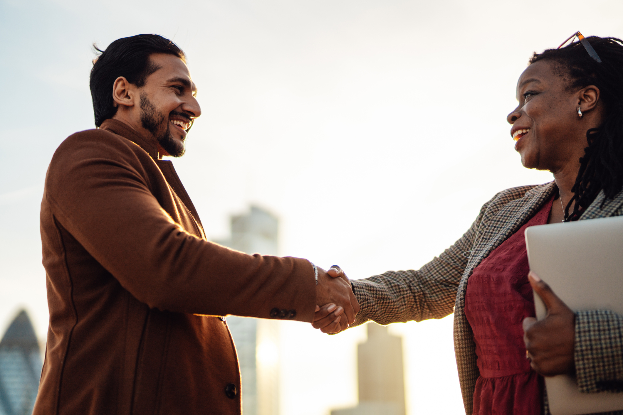 Two professionals shaking hands outdoors, one holding a laptop, both smiling