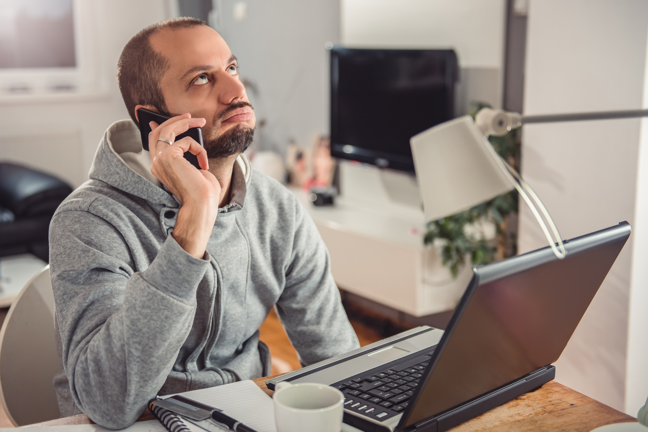 Person in a hoodie at a desk with a laptop, talking on the phone