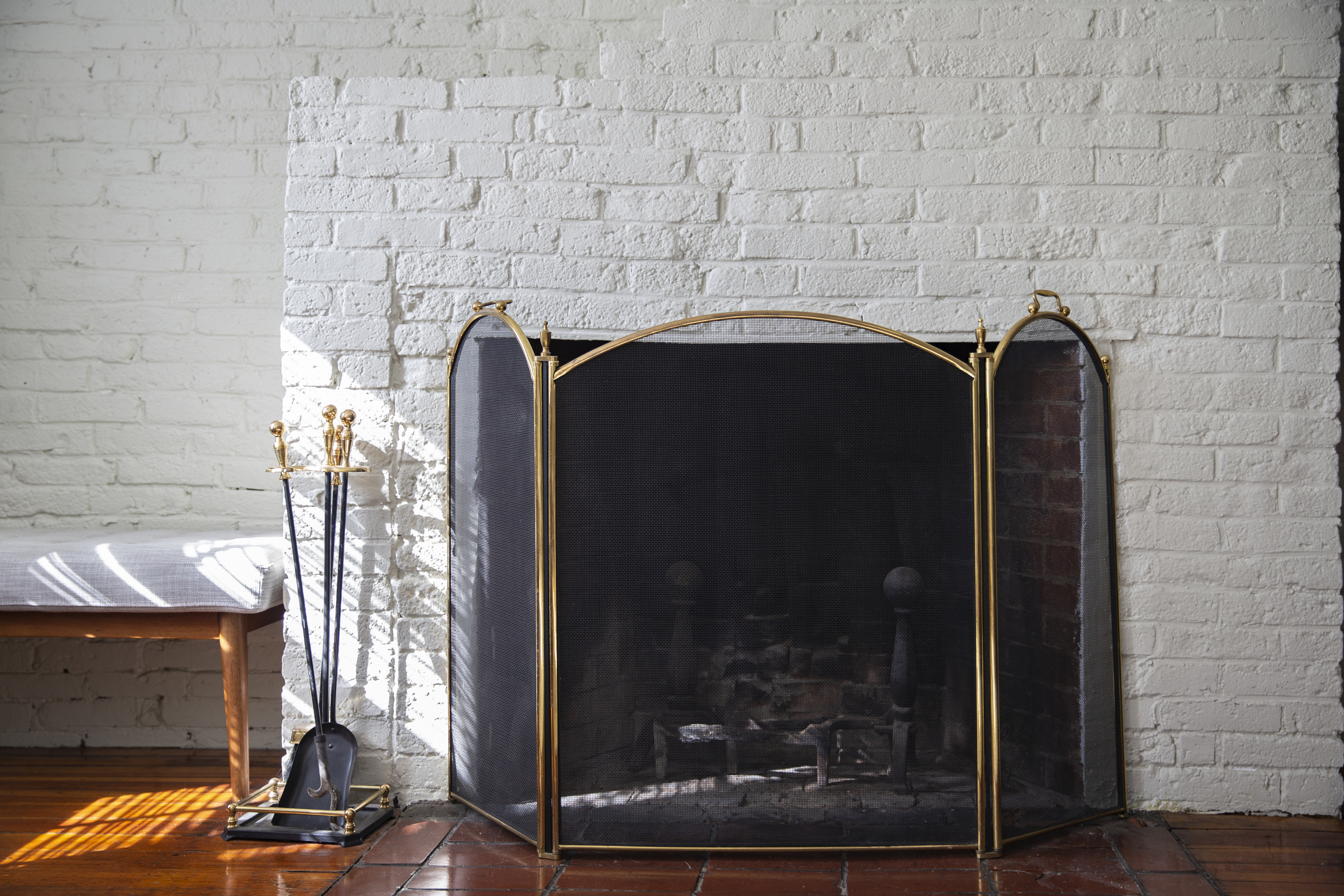 A fireplace with a mesh screen and tools against a white brick wall, next to a wood bench